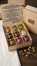 Handcrafted laser-engraved, fire branded birch wooden gift box open to reveal nine 1.5oz glass tasting jars of nut butters & nut spreads nestled in natural wood wool, displaying a spectrum of colorful artisanal nut butter spreads on a rustic wood table.