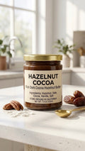 A glass jar of Hazelnut Cocoa Zero Added Sugar Spread on a sunlit marble counter, surrounded by whole roasted hazelnuts, raw cacao nibs, dates, and a gold spoon dripping with rich chocolate spread.