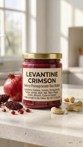 A glass jar of Levantine Crimson Pomegranate Nut Butter with a gold lid resting on a marble countertop, surrounded by fresh pomegranate seeds, sumac spice, and a golden spoon of deep crimson spread.