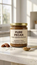 A glass jar of Pure Pecan Single-Ingredient Nut Butter with a gold metal lid on a sunlit counter, surrounded by whole raw pecans and a spoon of velvety amber pecan butter.