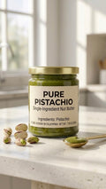 A glass jar of Pure Pistachio Single-Ingredient Nut Butter on a light countertop, flanked by vibrant raw pistachios and a golden spoon filled with stone-ground emerald green pistachio butter.