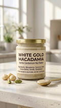 A glass jar of White Gold Macadamia Nut Butter with a gold lid on a stone countertop, framed by raw macadamia nuts, whole cardamom pods, vanilla bean pods, and a golden spoon of creamy ivory spread.